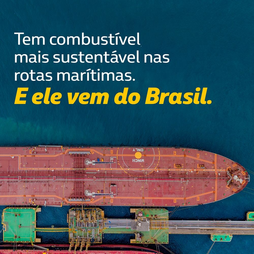: A imagem mostra uma vista aérea de um navio de carga, possivelmente um petroleiro, atracado em um porto. O navio é de cor vermelha, e há estruturas portuárias ao redor, com uma superfície aquática azul ao fundo. O texto inserido diz: "Tem combustível mais sustentável nas rotas marítimas. E ele vem do Brasil.