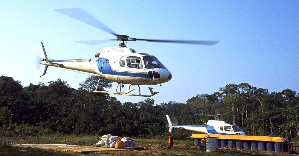 A imagem mostra um helicóptero em voo, com as lâminas girando visivelmente. O helicóptero é de cor branca com detalhes em azul e possui a inscrição "HELISOL". Abaixo, há uma área de terreno com sacos empilhados, possivelmente de materiais ou suprimentos. Ao fundo, pode-se ver uma vegetação densa, típica de uma área florestal. À direita, há outro helicóptero estacionado.