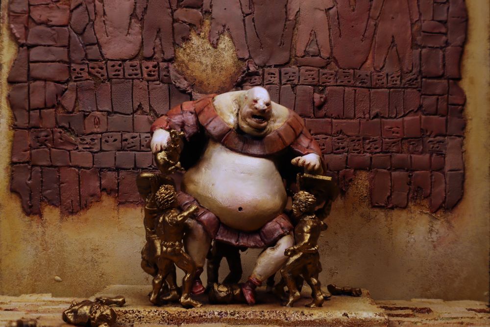 Closeup of a diorama of a portly tyrant sitting on a golden throne in front of a wall of hanging flesh. 