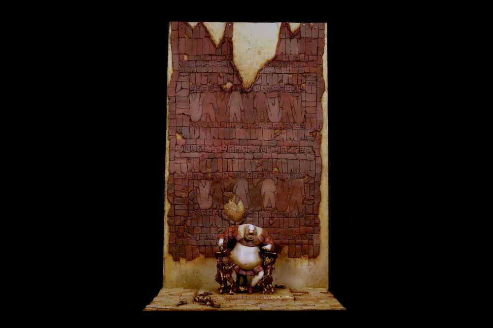 Diorama of a portly tyrant sitting on a golden throne in front of a wall of hanging flesh. 