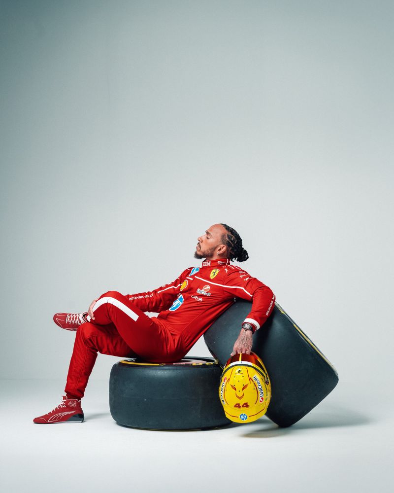 Photo of 7 time F1 world champion Sir Lewis Hamilton in his Ferrari racing suit, sitting on a pair of tires while holding his helmet. 