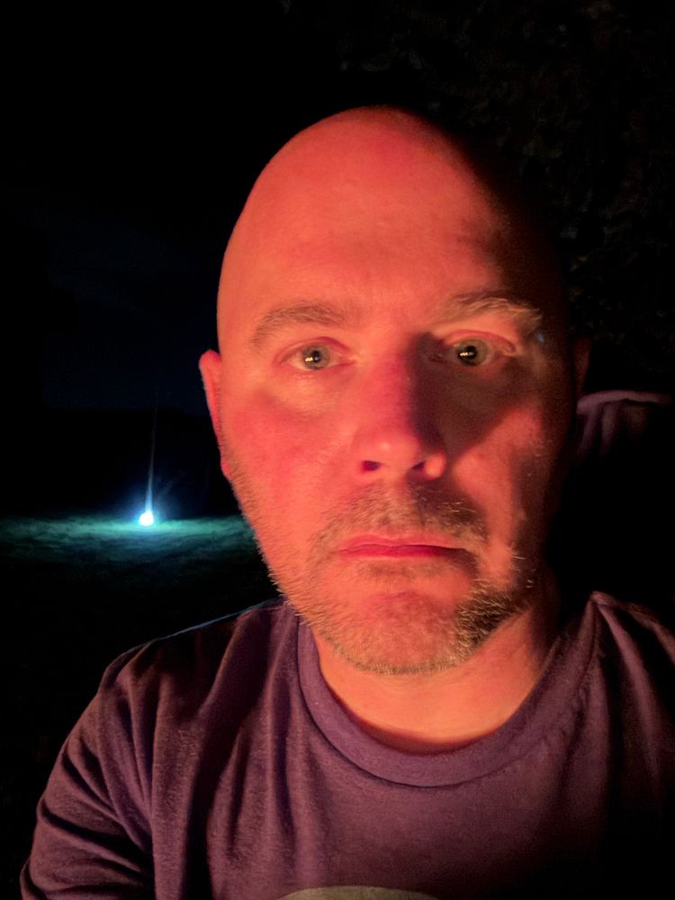 A middle-aged bald man staring at the camera and the fucking void, his face lit by the fire. 