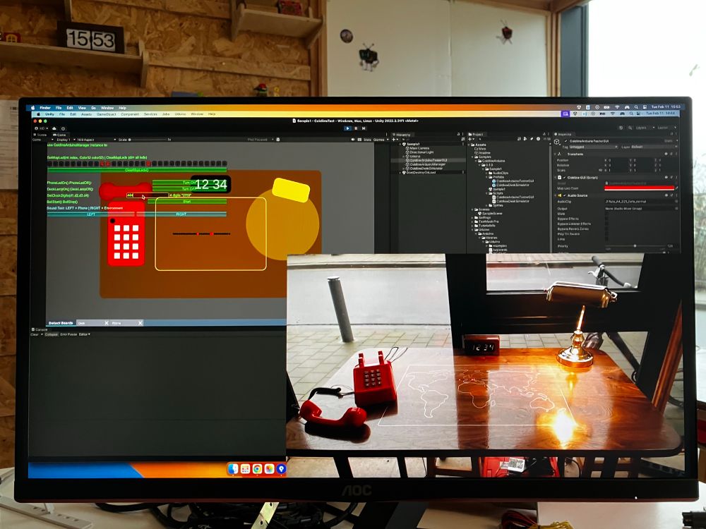 A monitor showing game engine software and a webcam feed of an interactive art installation with a desk and a phone.