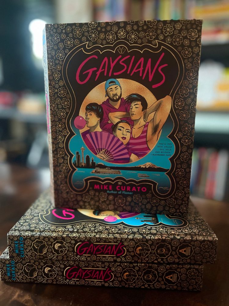 A photo of the book Gaysians by Mike Curato prominently displayed upright on a table, with two additional copies stacked horizontally beneath it. The cover features vibrant, stylized illustrations of five queer Asian individuals in dynamic poses, surrounded by a floral pattern with a gold and black border. The title "Gaysians" is written in bold, hot pink letters. The background includes an illustrated skyline and Mount Rainier, symbolizing a Pacific Northwest setting. The spine of the book shows the title, author’s name, and decorative floral motifs. The image is taken in a bookstore or library setting, with shelves of books blurred in the background.