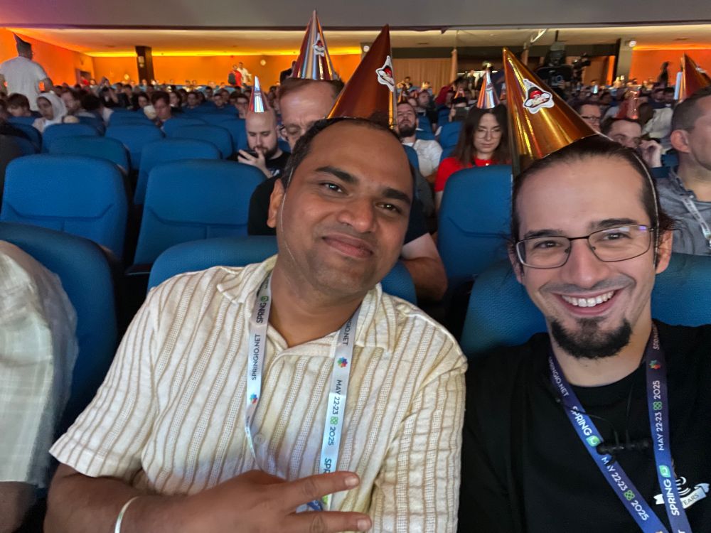 Selfie of Daniel and Bala at Spring I/O, wearing golden party hats with a Duke sticker on it.
They are smiling.
People behind them are also wearing the silly cone hat.