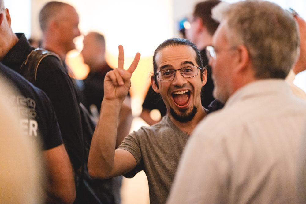 Daniel, still in the same conversation, looking at the camera, making an ecstatic V-sign with his fingers, huge smile on his face