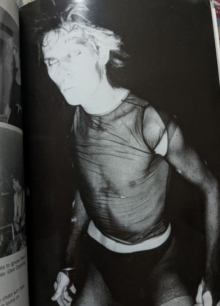 Freakish skinny androgenous white man with wild hair wearing sheer tight black top, his face partially whited out by bright light