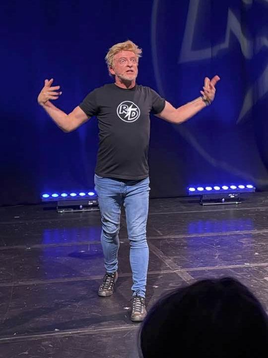 Rhys Darby onstage at the Edinburgh Fringe in his RD tshirt and blue jeans. His eyes are wide open in a slightly manic look. 