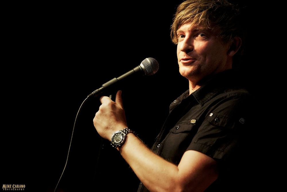 Rhys Darby on stage with a mic, wearing a short sleeved shirt fighting for its life against his naturally big biceps