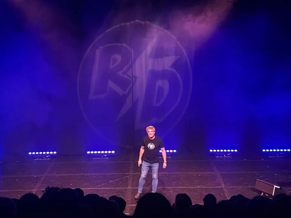 Rhys Darby on stage at the Edinburgh Fringe in his The Legend Returns show. The picture is taken from quite far back in the audience , making him appear quite small. He appears to have an aggressive stance and expression.
