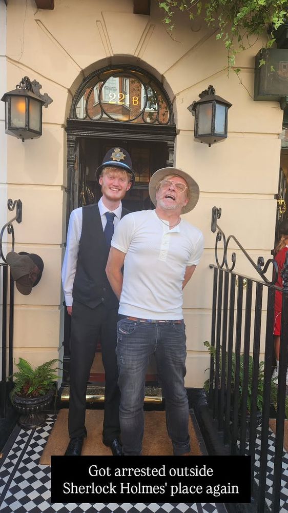 Picture from Rhys Darby's IG of him outside 221B Baker Street in London being "arrested" by the doorman who is wearing an English policeman's hat.. The caption says "Got arrested oustide Sherlock Holmes' place again"