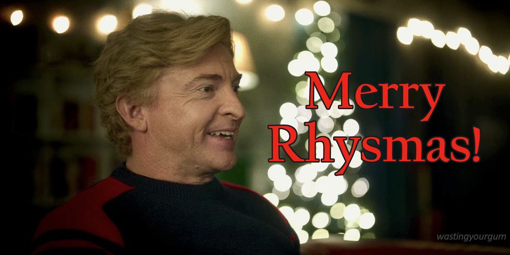 Rhys Darby as Casper in "Relax, I'm From the Future". He's wearing a red and blue sweater and smiling at something over to the right. There is a Christmas tree with white lights out of focus in the background behind him.
