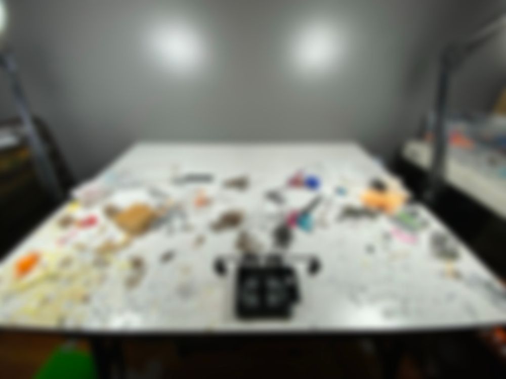 a blurred image of Loft Studios desk, it is entirely messy. random yellow scraps are on the left corner as a orange repo character lies there. a handmade lego phone holder stands in the middle. hazard(?) and sarah are also seen in the middle of the desk based on their colors.