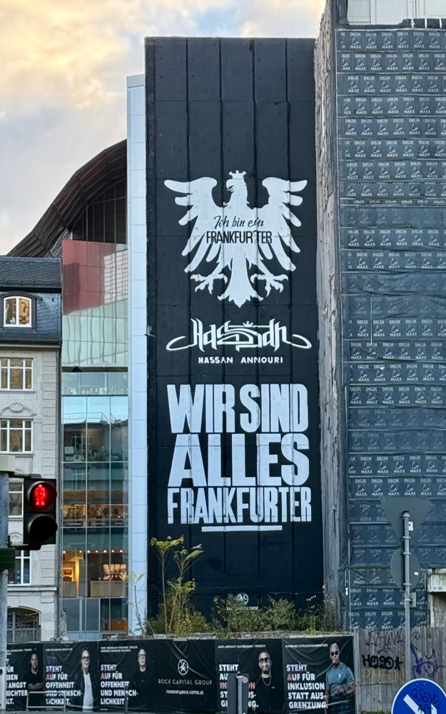 Hauswand, die schwarz gestrichen ist. Darauf ist in weiß ein stilisierter Frankfurter Adler gemalt in dem in Schreibschrift „Ich bin ein Frankfurter“ steht. Darunter steht etwas in arabischer Schrift, darunter klein Hassan Annouri und darunter in groß „Wir sind alles Frankfurter“. 