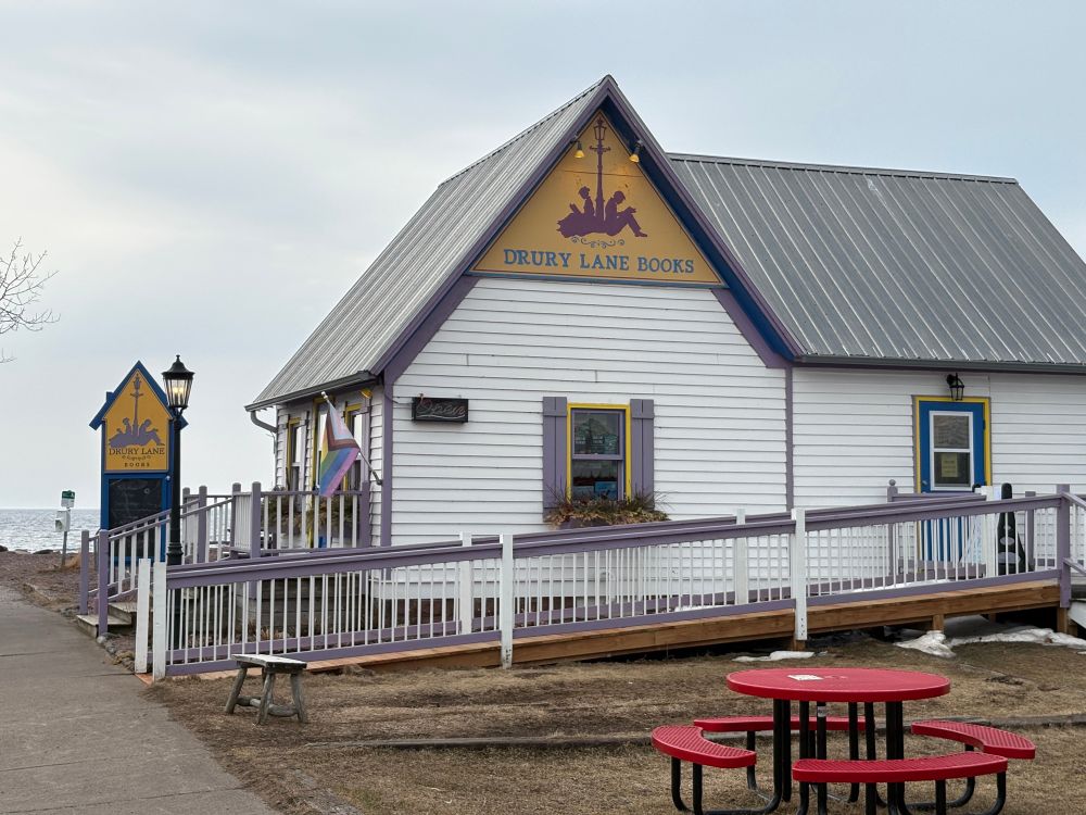 Drury Lane Books store. White sided house with a purple and ramp outside. Drury Lane Books at the roof peak in yellow and purple. 