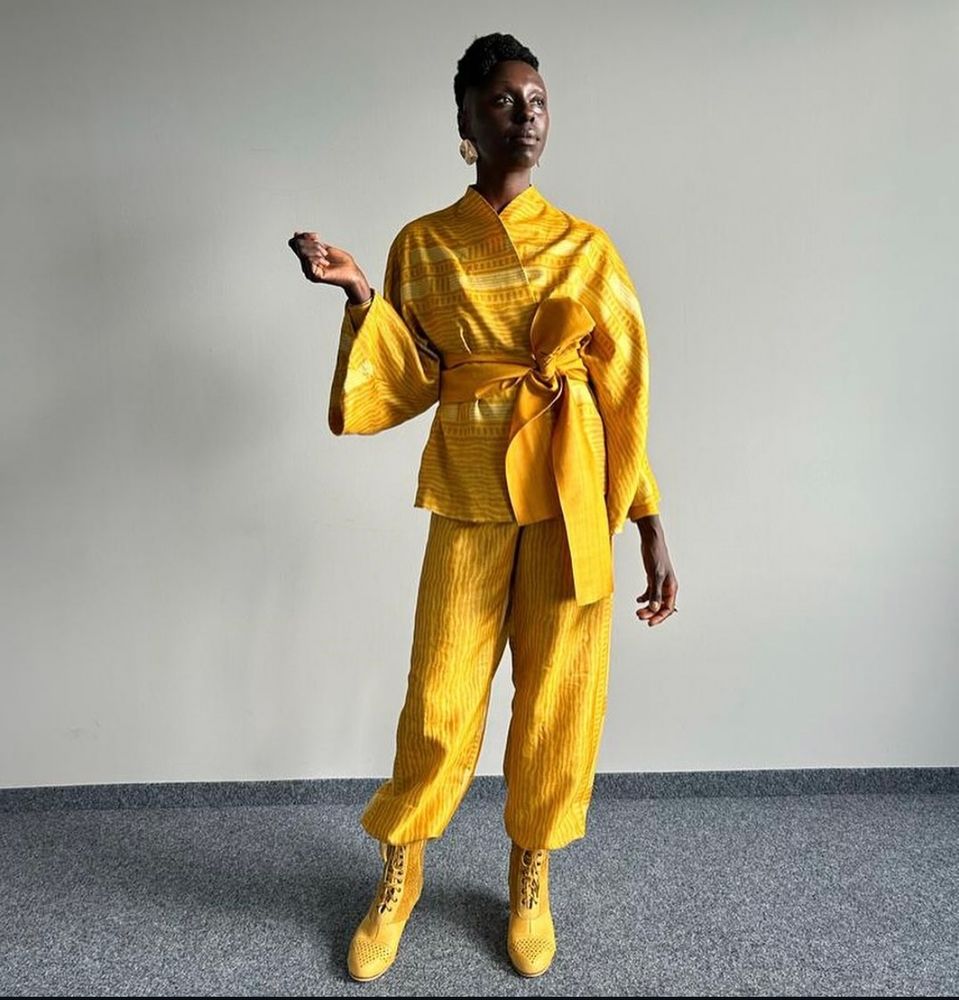 Actress Nyokabi Gethaiga plays Ryma Sedai of the Yellow Ajah. Dressed all in yellow in a long wide sleeved top with high collar and tied around her waist with a wrapped yellow length of material. Yellow pants and boots. 