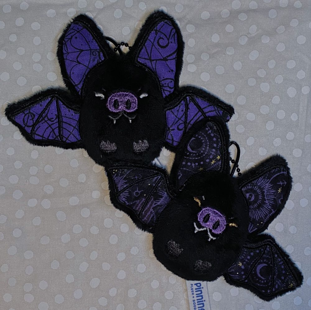Two bat keychain plush, black with grey and purple details. One has spiderweb pattern and the other has a general occult/astrological pattern.