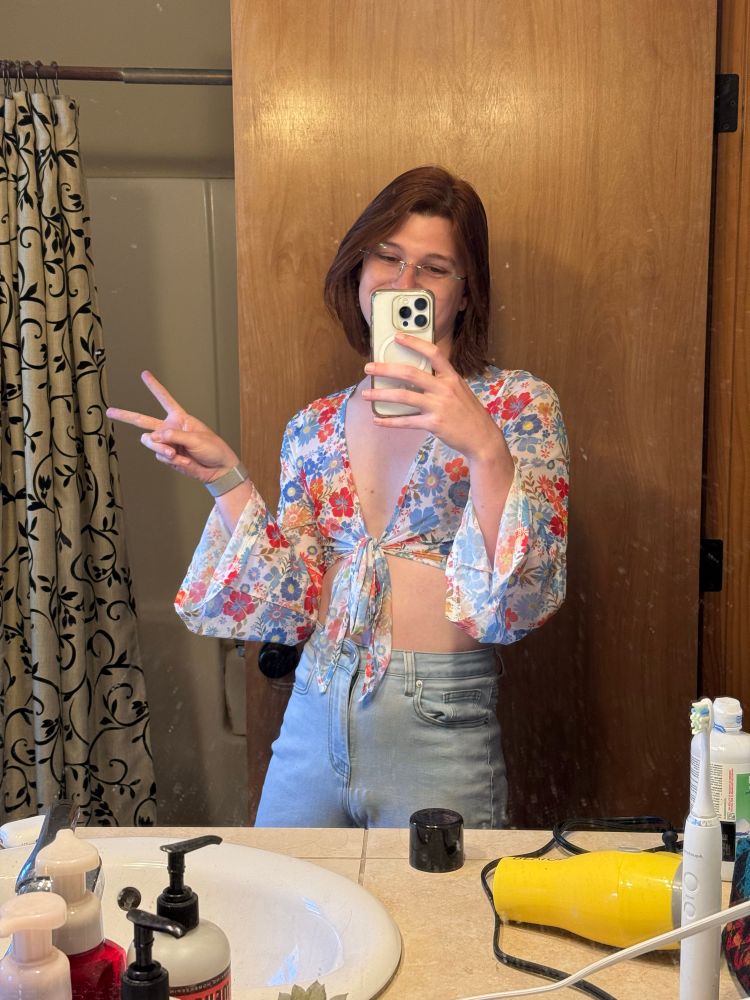 A woman poses for a mirror selfie holding up a peace sign. She is wearing a blue and red flower pattern crop top that ties in the middle with long sleeves. 