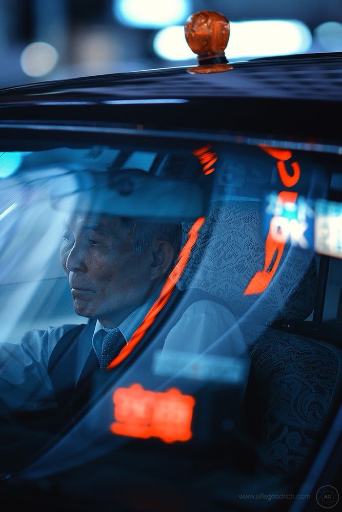 A taxi driver sat waiting for the traffic lights at Tokyo’s famous Shibuya Crossing. It’s night and the blue hues of the a cool white balance on the camera contrast with the reds of the sign in his cab that says he’s taking fares. And the reds of reflected neon signs above. 