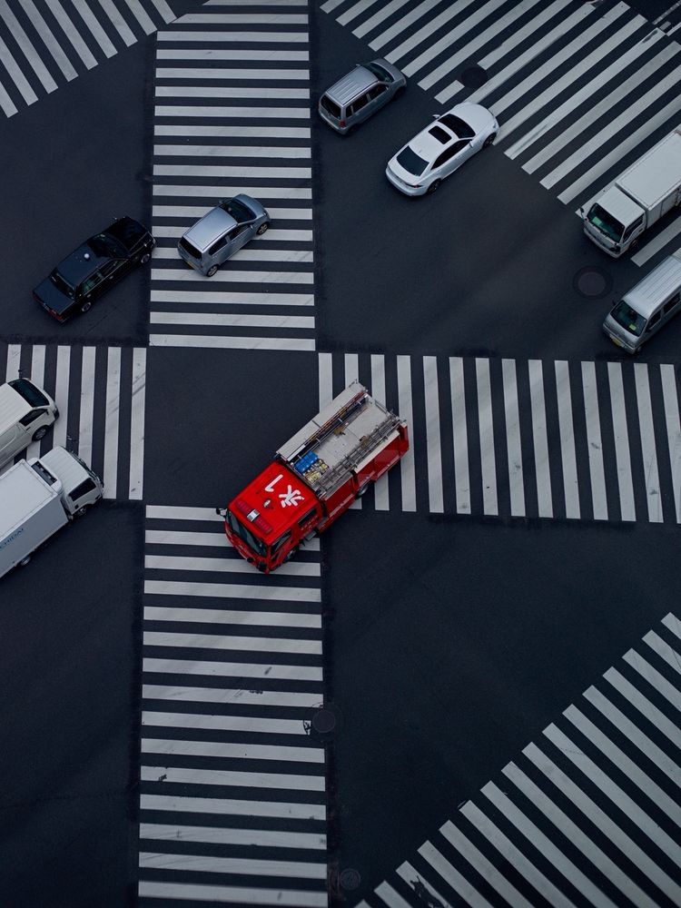 A view down on to the large zebra crossing in Tokyo’s Ginza district. White, silver and black cars pass through. In the centre is red fire engine. 