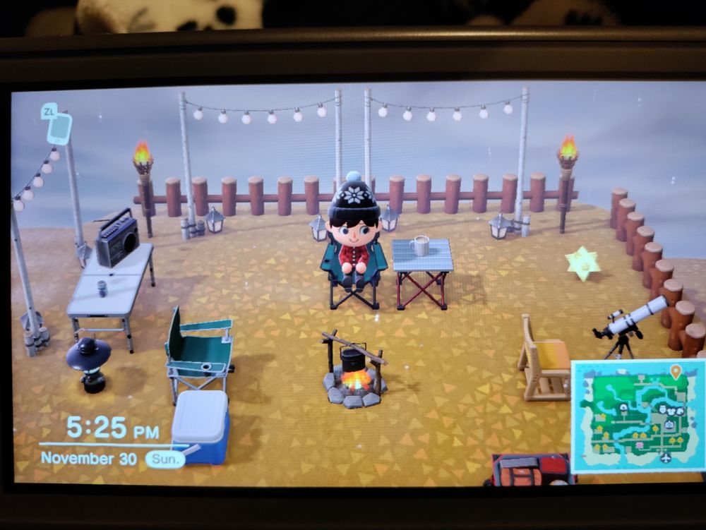 A photo of a cozy, star-gazing campsite in Animal Crossing: New Horizons, with the player sitting in the middle. It has lights, chairs, a cooking pot over the campire, a music player, a cooler for refreshments and a telescope. Perfect for friends to visit and hang out 