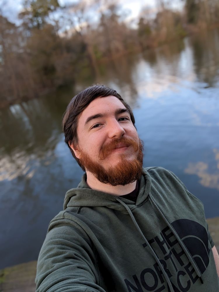 Picture showing Alex against a slightly blurred lake with trees showing on the opposite shore. The sunlight is showing a nice contrast between his dark brown hair and red beard.