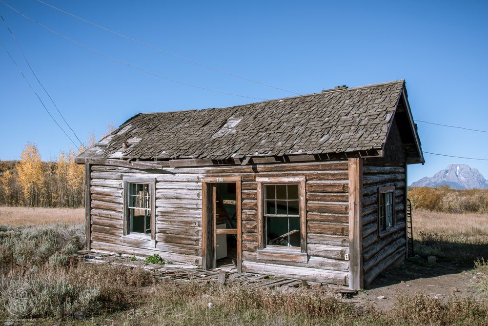A color photograph of an old log cabin with a collapsing wooden shingle roof and broken windows sitting alone in a grassy field. The door hangs ajar, revealing part of the abandoned interior. Autumn trees line the horizon behind the cabin, and a snow-capped mountain peak from the Teton Range rises in the far distance under a bright blue sky.