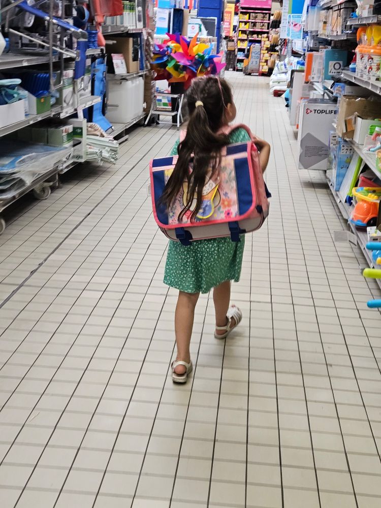 Petite fille en robe verte et cartable rose et bleu sur les épaules. 