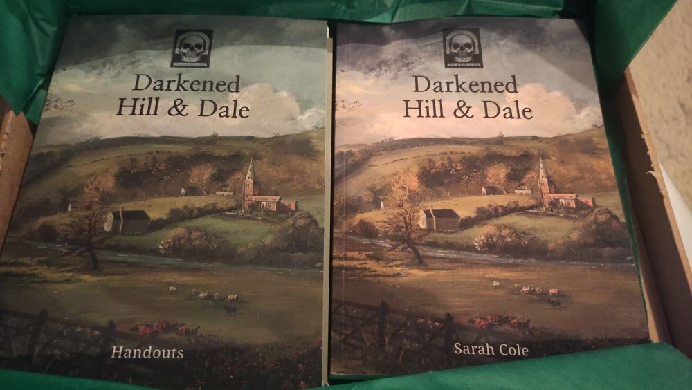 An unboxing for Darkened Hill and Dale with the main booklet on the right and handouts on the left. Both have a bucolic rural scene on the front and a "this blighted Isle" sticker with a skull on it 