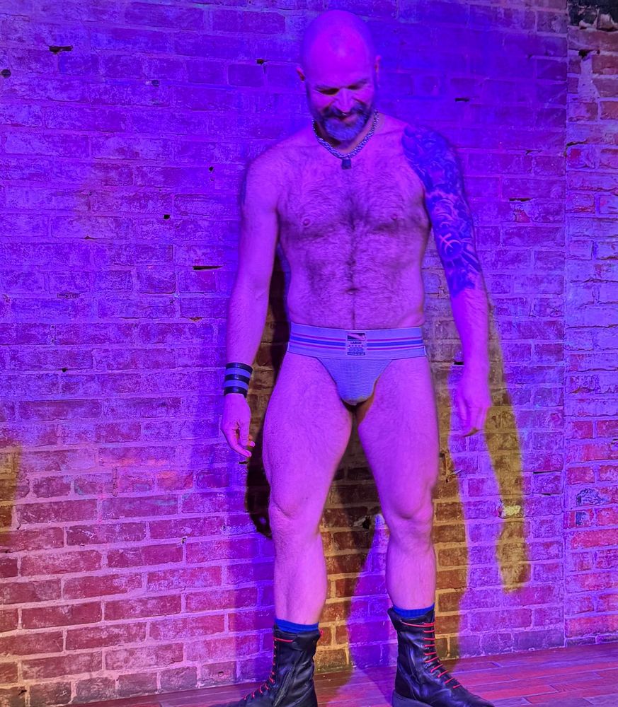Man in jockstrap and boots stands shyly on stage at a gay bar