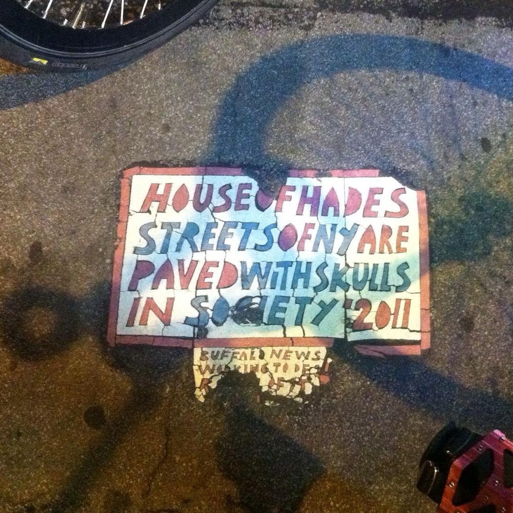 A worn looking white vinyl tile with faded red and blue lettering embedded in some greasy looking asphalt. The lettering is hand cut and the tile is cracked. It reads “Hoise of Hades Streets Of NY Are Paved With Skulls In Society ‘2011”
Below that in smaller letters it says something like “Buffalo News Working to De…” but beyond that it is worn away and illegible.
