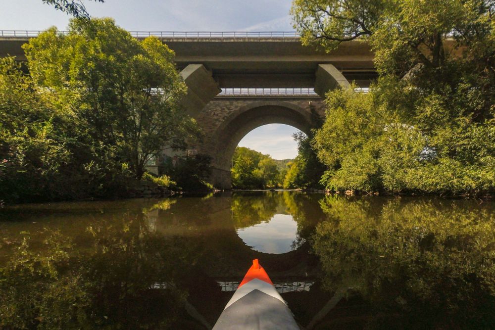 Blick aus einem Kajak auf einem ruhigen Fluss, der unter einer großen Steinbogenbrücke und einer darüber verlaufenden modernen Straßenbrücke hindurchführt, umgeben von dichtem, grünem Laub und Spiegelungen im Wasser.
