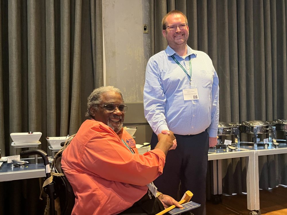 A man, standing, shakes the hand of a man, sitting, who's golding a gavel.