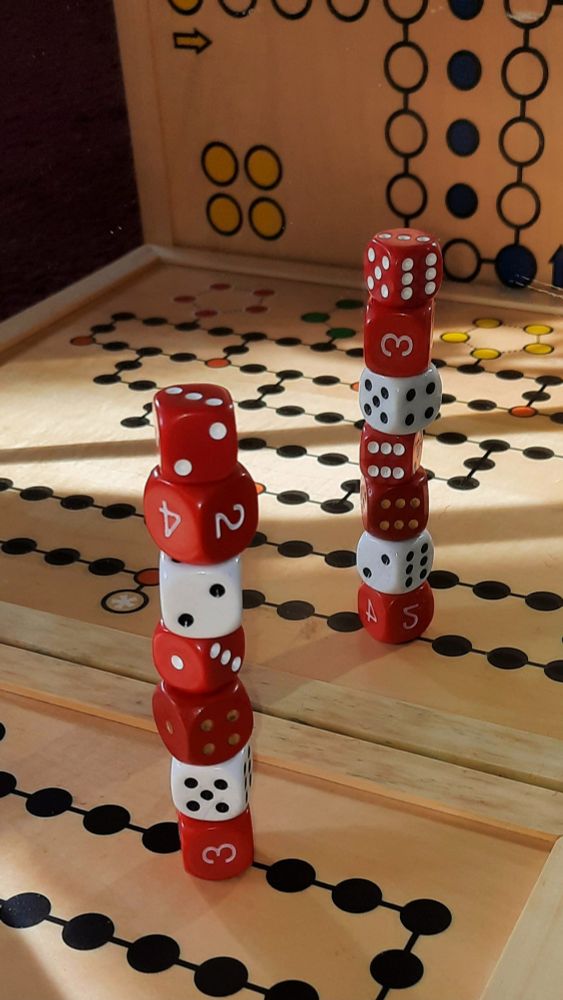 Ein Spielbrett Malefiz, im 90 Grad Winkel senkrecht dahinter ein Spielbrett Mensch-ärgere-dich-nicht. Darauf übereinander gestapelt 7 unterschiedliche, weiße und rote Spielwürfel. Das ganze vor einem Spiegel, so dass wir die gleiche Szenerie noch einmal sehen.