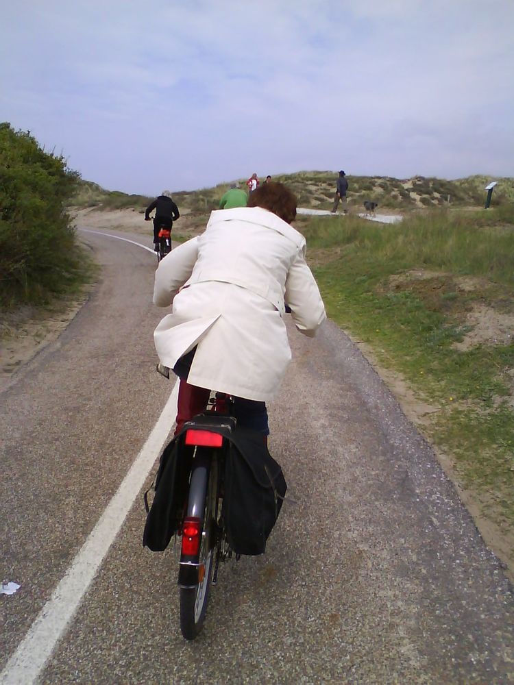 Leicht ansteigende Linkskurve auf dem Radweg durch die Dünen. Die Radlerin direkt vor uns kämpft sich hinauf. Ihr heller Mantel wird durch den Gegenwind aufgebläht.