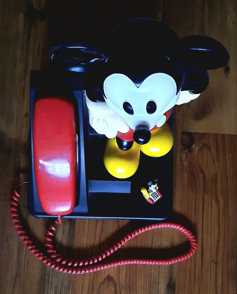 Ein großes, echtes Micky-Maus-Telefon mit rotem Hörer, senkrecht von oben fotografiert. Auf dem Sockel eine kleine Micky-Maus-Figur als Telefon mit gelbem Hörer.