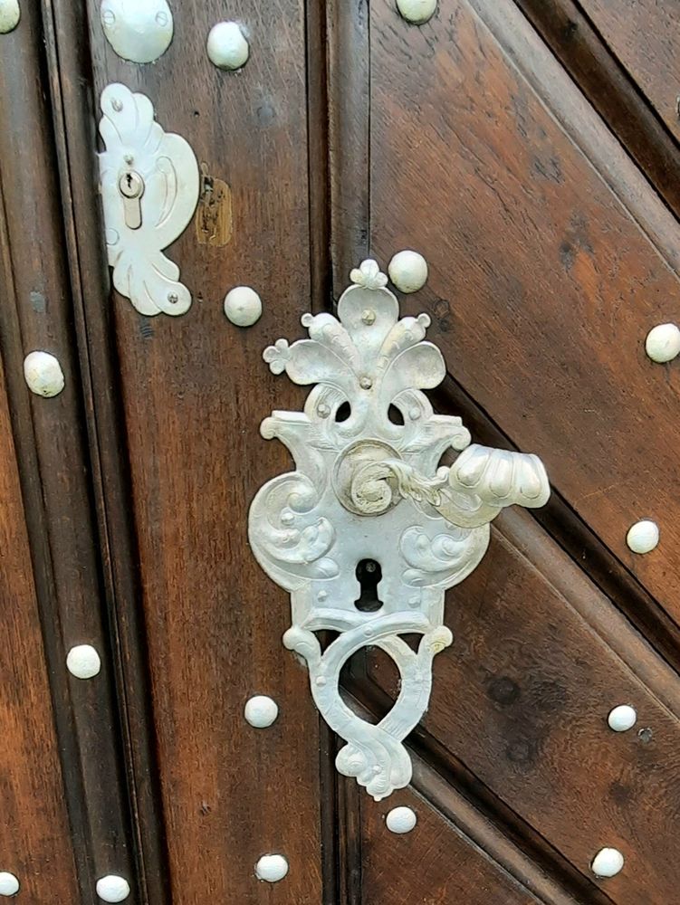 Eine dunkelbraune zweiflügelige Haustür. Sie ist von oben bis unten mit weißen Metallnieten besetzt. Dazu ein reich reich verzierter und verschnörkelter weißer Türgriff.