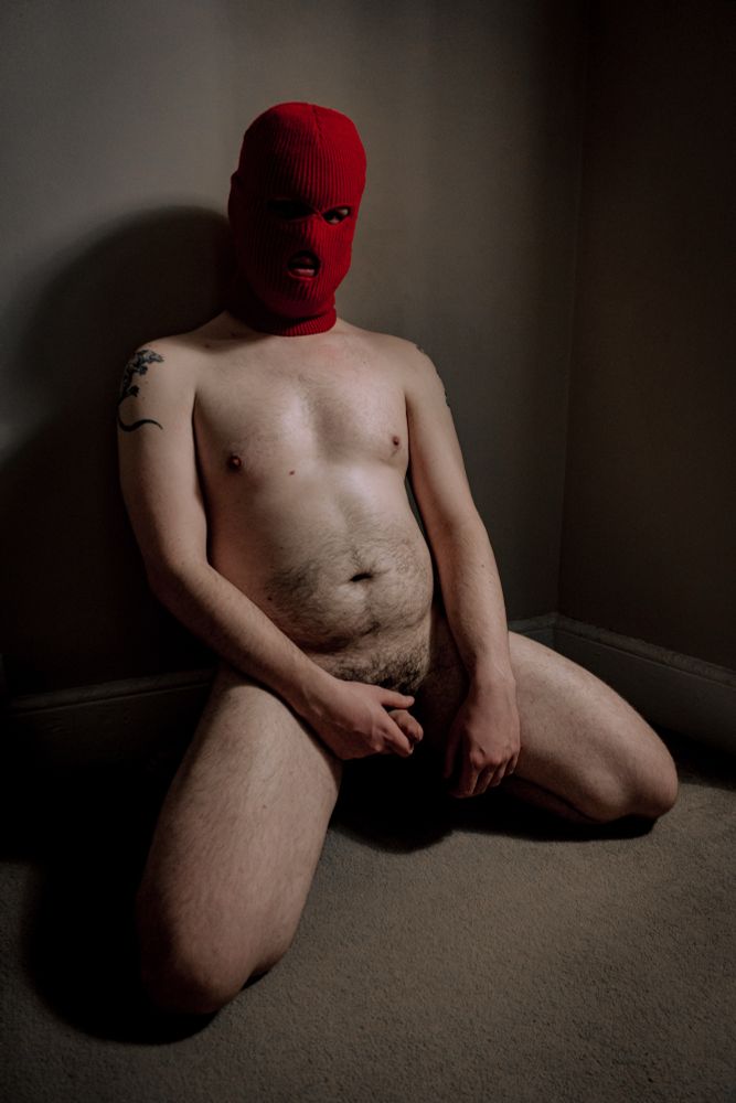 A naked man in a red balaclava kneels legs spread on the floor holding his cock.