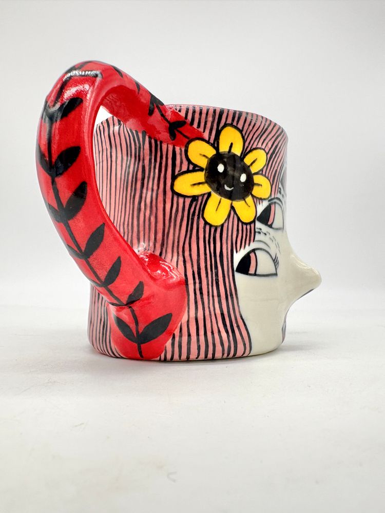 Side view of a slab-built, handpainted mug with a woman’s face with a 3d nose, pink hair, and a happy little flower. 