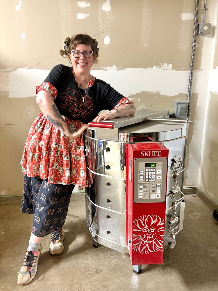 Middle aged white lady with tattoos wearing a black tshirt, red dress, black pants, and sneakers standing proudly next to her brand spanking new skutt 1027 kiln. 