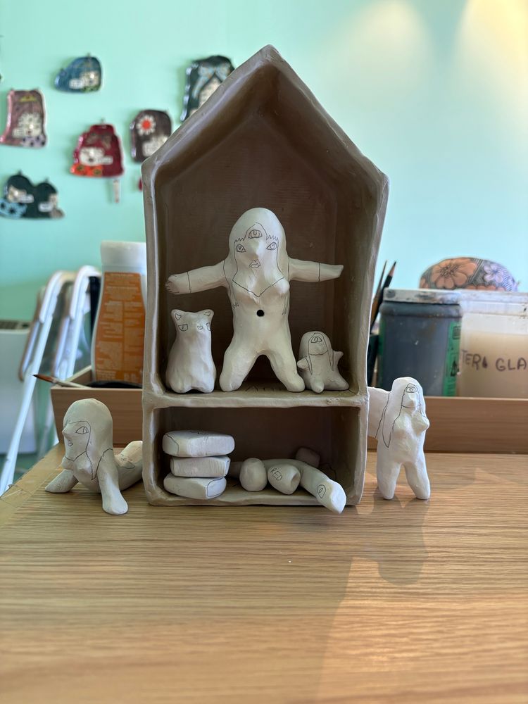 Ceramic greenware house with a woman, baby, and cat on the upper floor. Beneath them are books, apples, and a snake. Parade are two figures. 