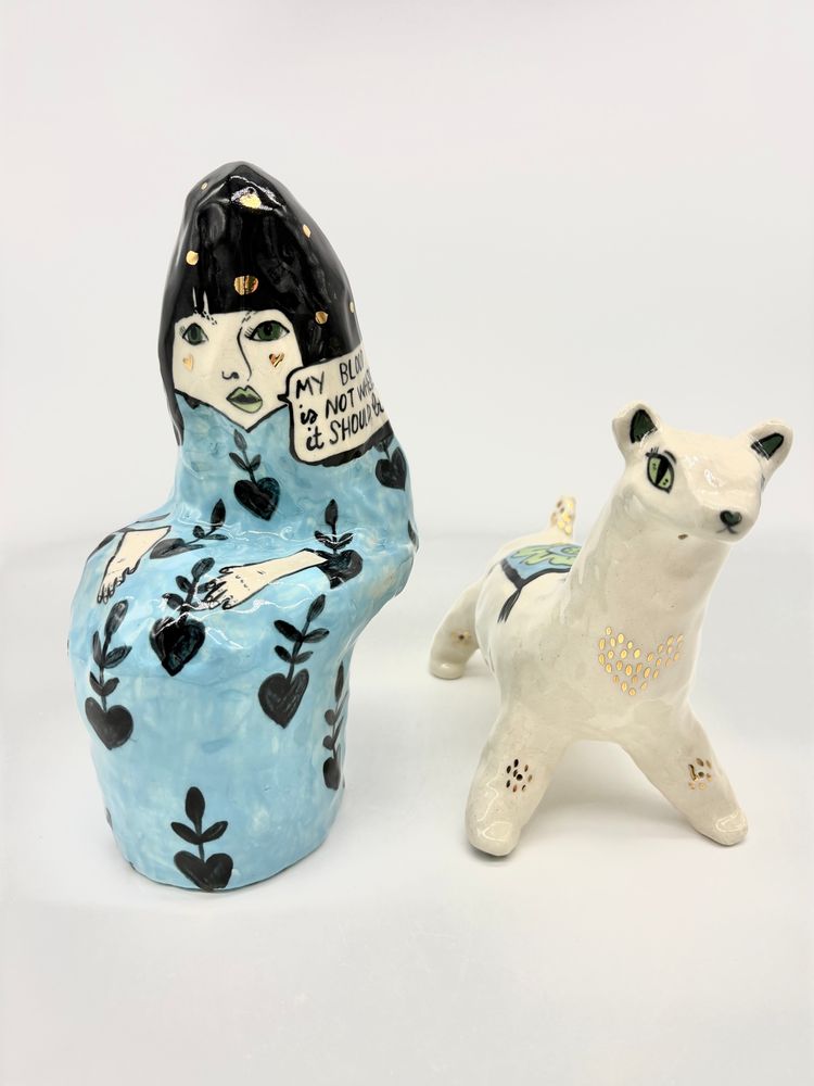 A set of ceramics featuring a seated woman in a blue dress and a white weasel that is her companion. They are spaced slightly apart. 