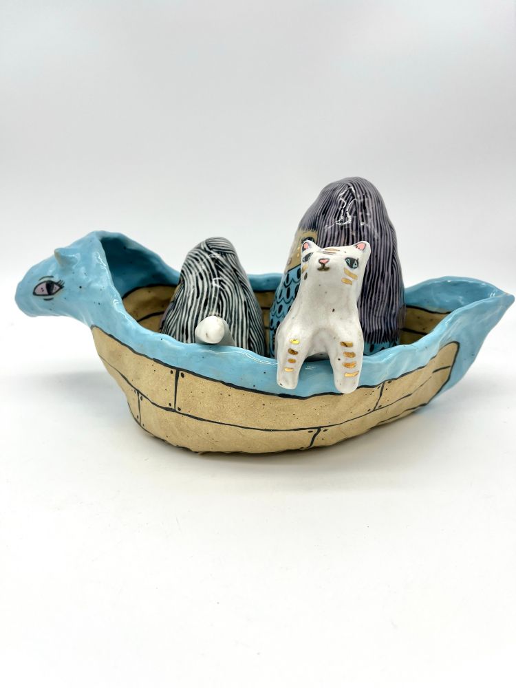 A ceramic boat with an animal head that carries two ceramic women and a ceramic serpent cat. Side view with cat