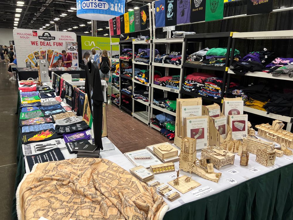 A booth at Origins Game Fair 2025 with tables containing various nerdy t-shirts, stickers and laser cut toys for the undiscerning gamer. In the back of the booth has shelves full of t-shirts.