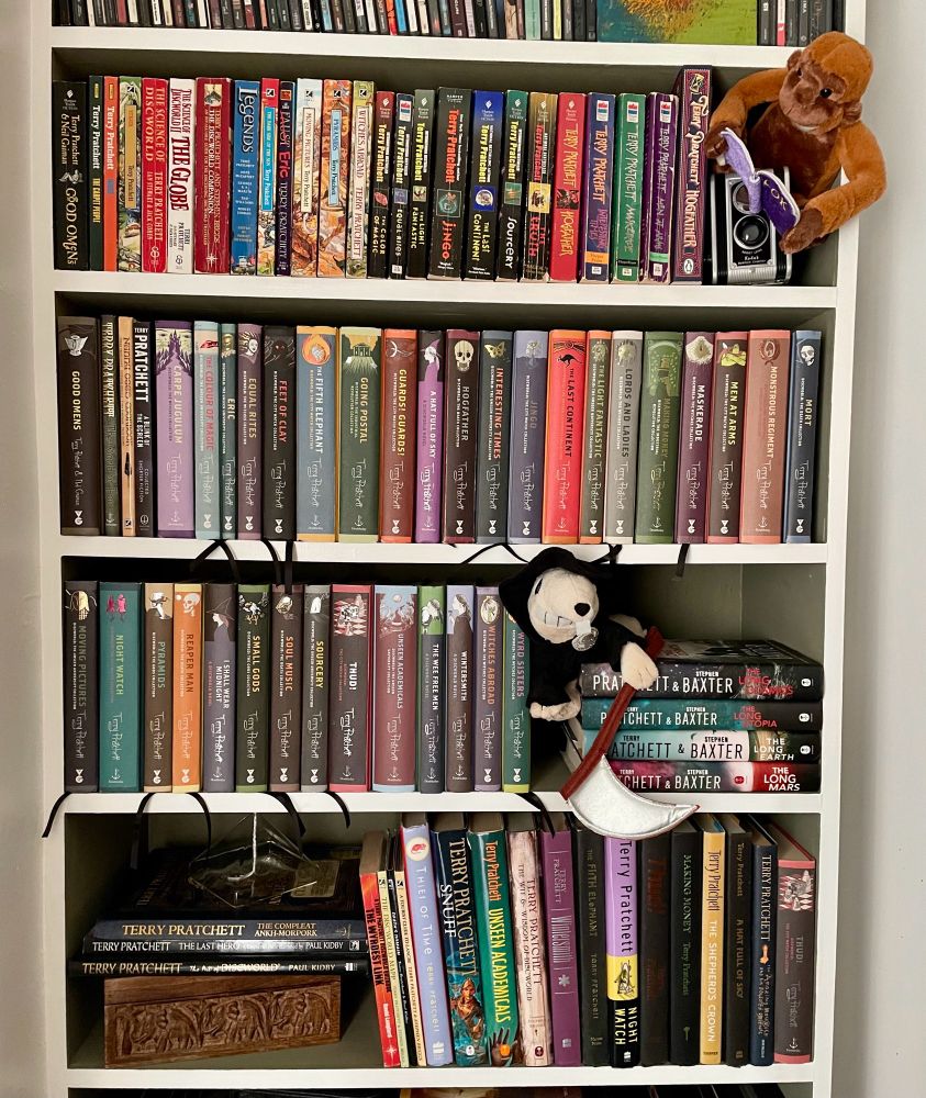 Four shelves of Terry Pratchett books, including lots of Discworld. A Librarian plush toy holds a book that says ook and sits on an old camera in a nod to Otto Chriek. A plush Death of Rats peeks out from a shelf. A glass pyramid sits atop a wood box with carved elephants that holds a Thud game.