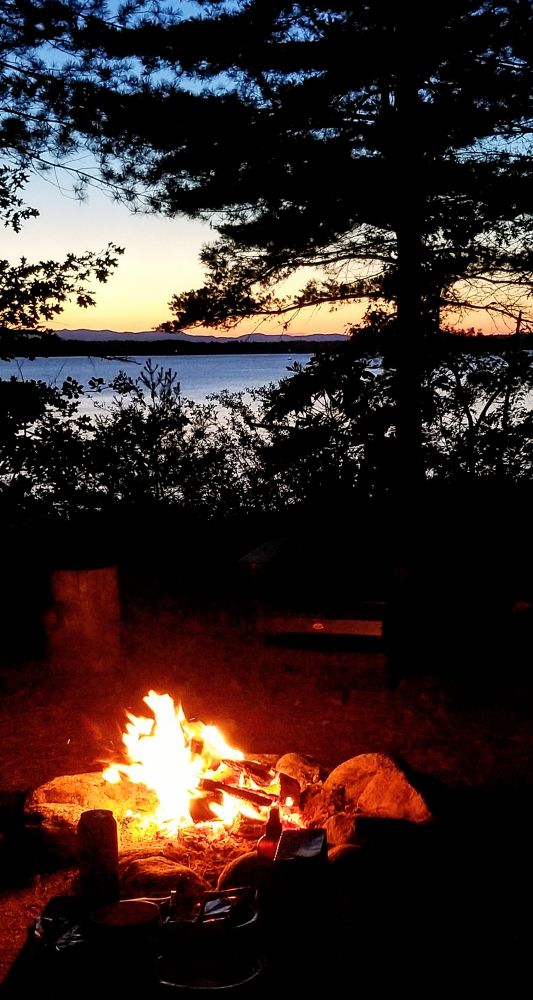 A campfire is in the foreground.  A lake, mountains, and sunset in the distance with pine trees silhouetted.