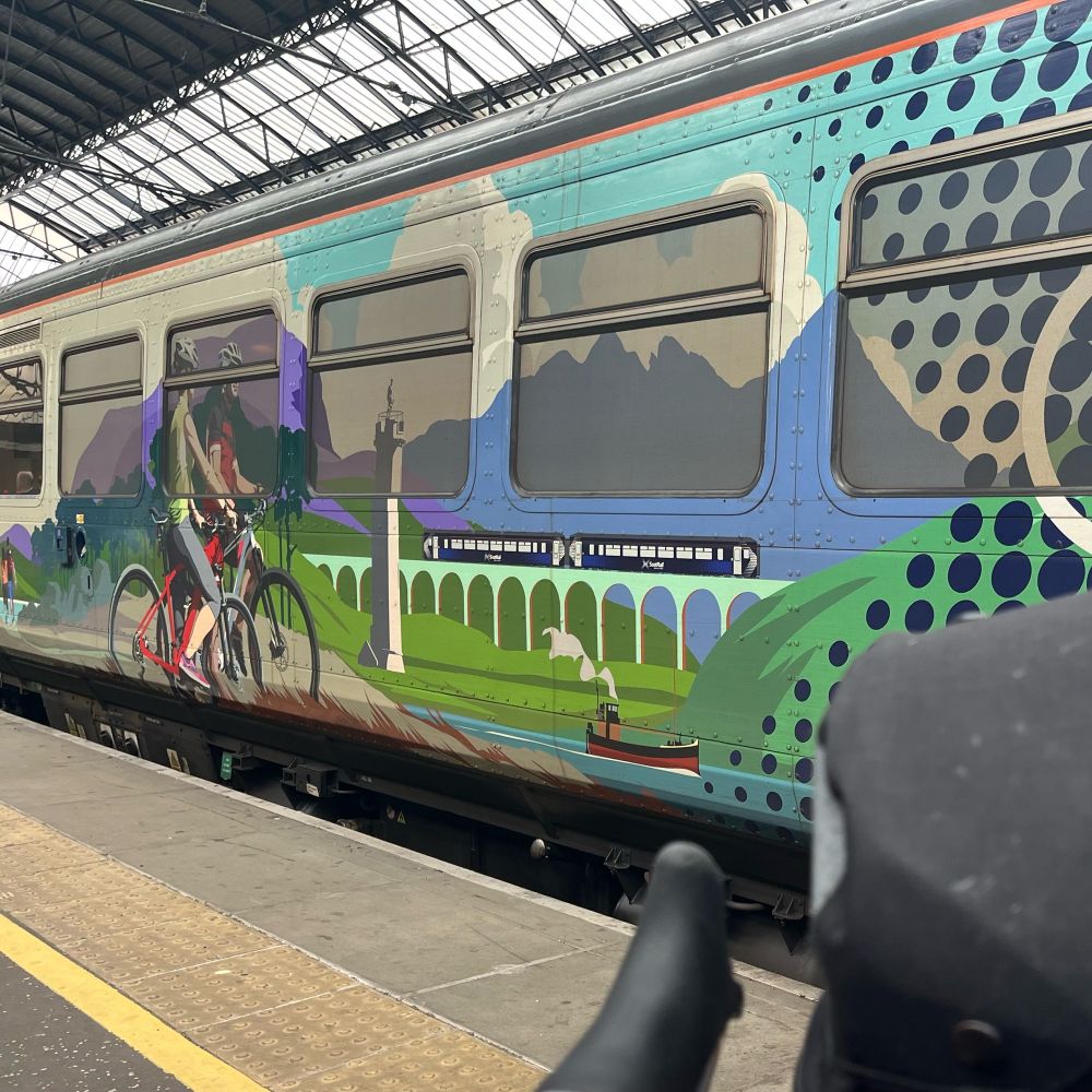 The handlebars of a bike in front of a train carriage. The carriage features an image of cyclists along with Scottish scenery 🏴󠁧󠁢󠁳󠁣󠁴󠁿 