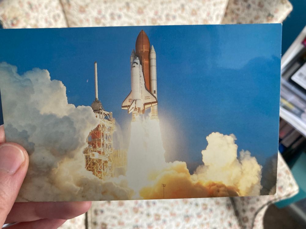 A photo of the Challenger space shuttle lifting off. There is a white and orange rocket rising into a cloudless blue sky with plumes of smoke behind it 