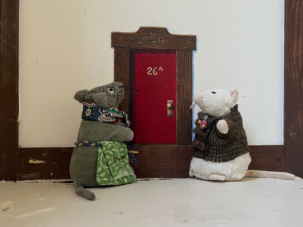 Two plush mice standing in front of a red mouse-sized door painted on the side of a fireplace. 