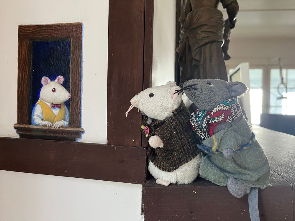 Two plush mice standing on the edge of a fireplace mantle looking at painting of a similar mouse leaning towards them from a window. 